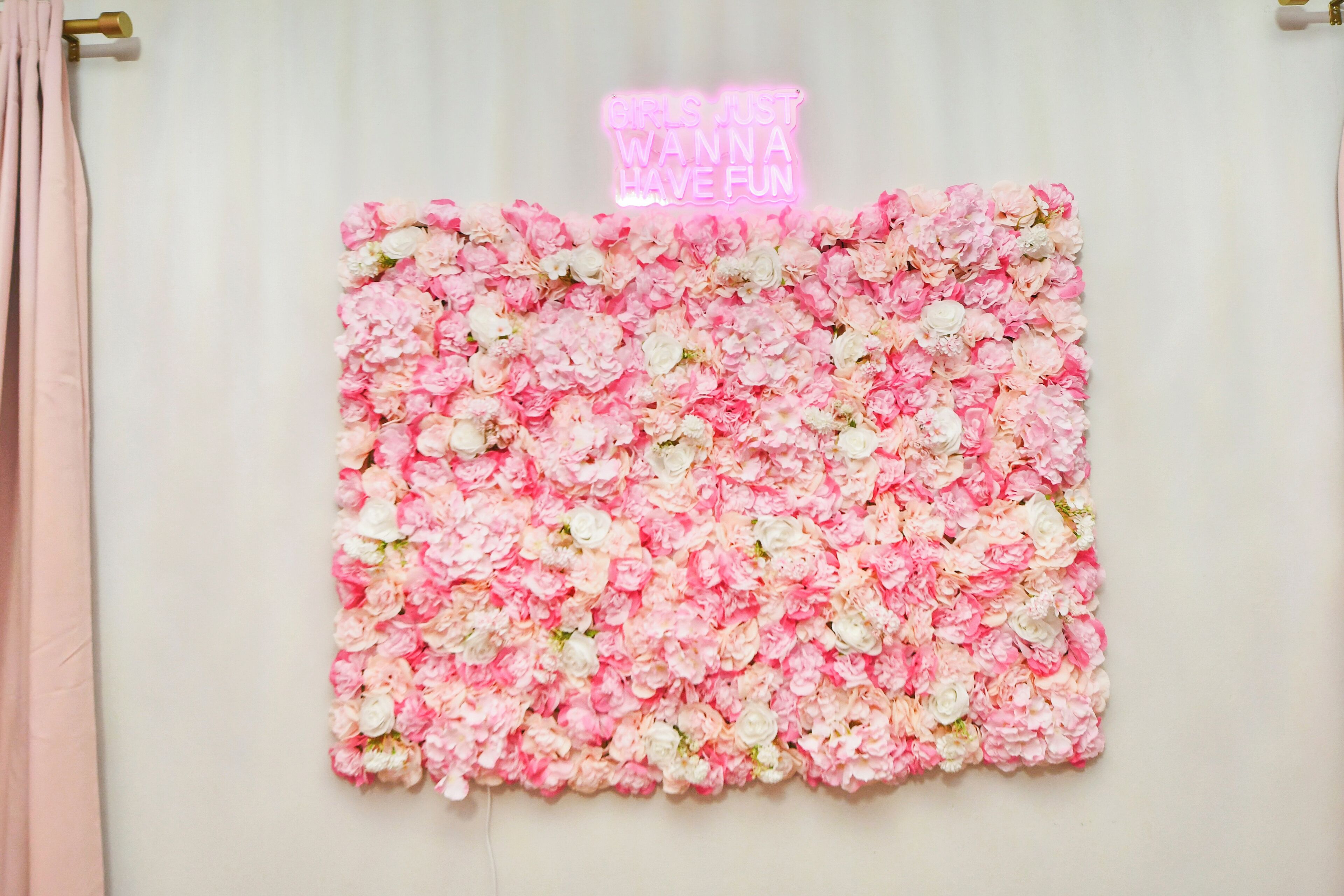 A wall covered with pink and white flowers and a neon sign that reads "Girls Just Wanna Have Fun."