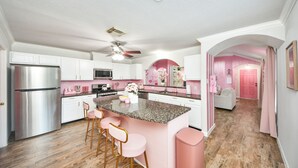 A modern kitchen with white cabinets, a stainless steel refrigerator, pink bar stools, and a granite countertop island.