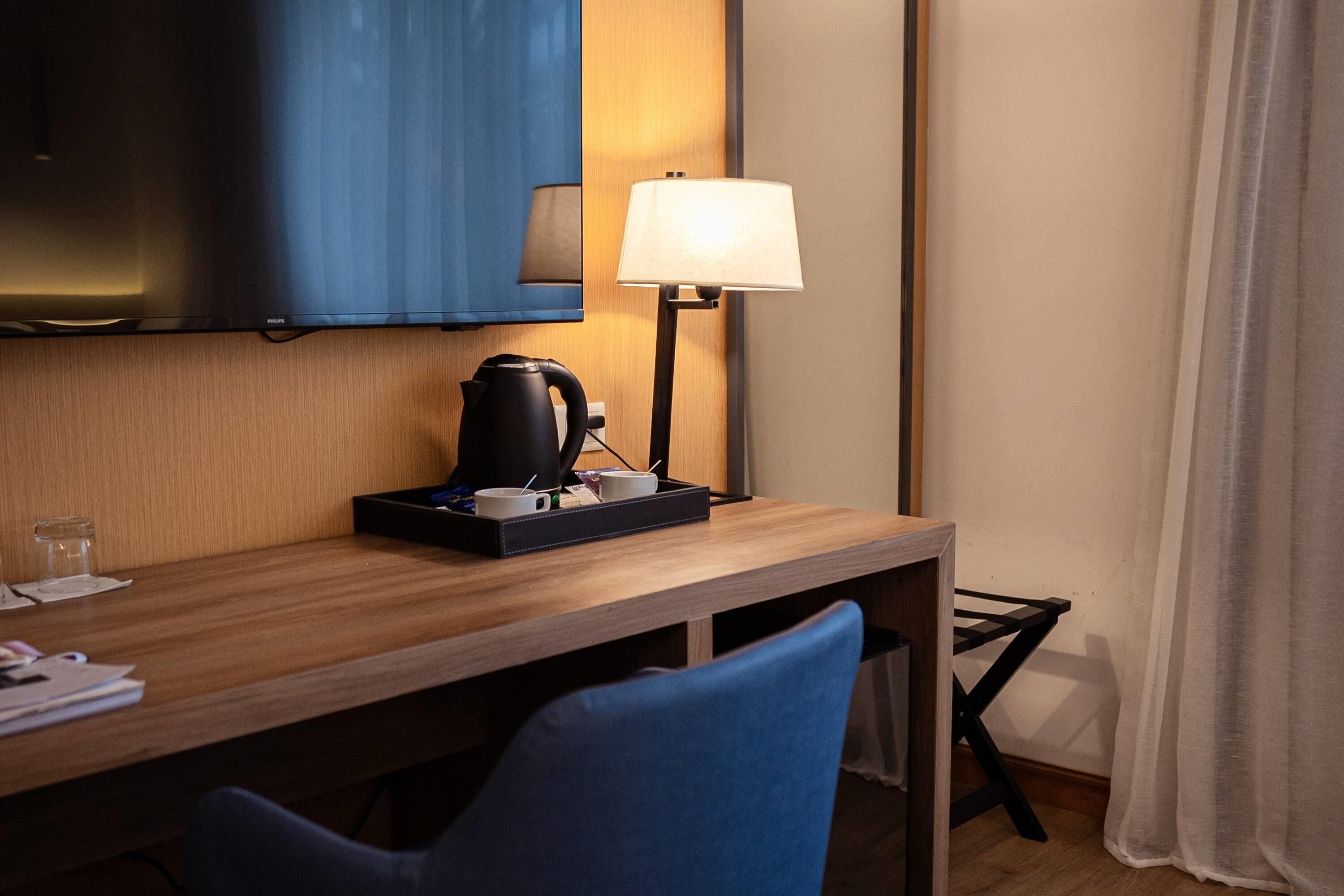 A hotel room with a wooden desk, a black kettle, a white mug, a blue chair, a television mounted on the wall, and a bedside lamp.