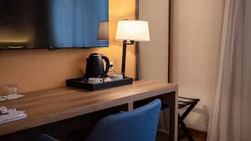 A hotel room with a wooden desk, a black kettle, a white mug, a blue chair, a television mounted on the wall, and a bedside lamp.