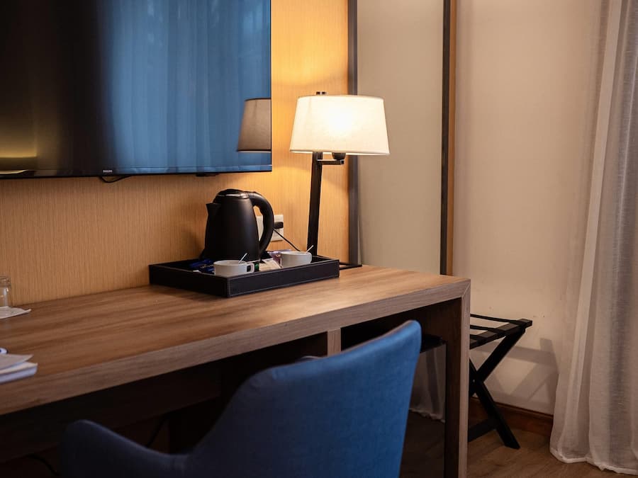 A hotel room with a wooden desk, a black kettle, a white mug, a blue chair, a television mounted on the wall, and a bedside lamp.