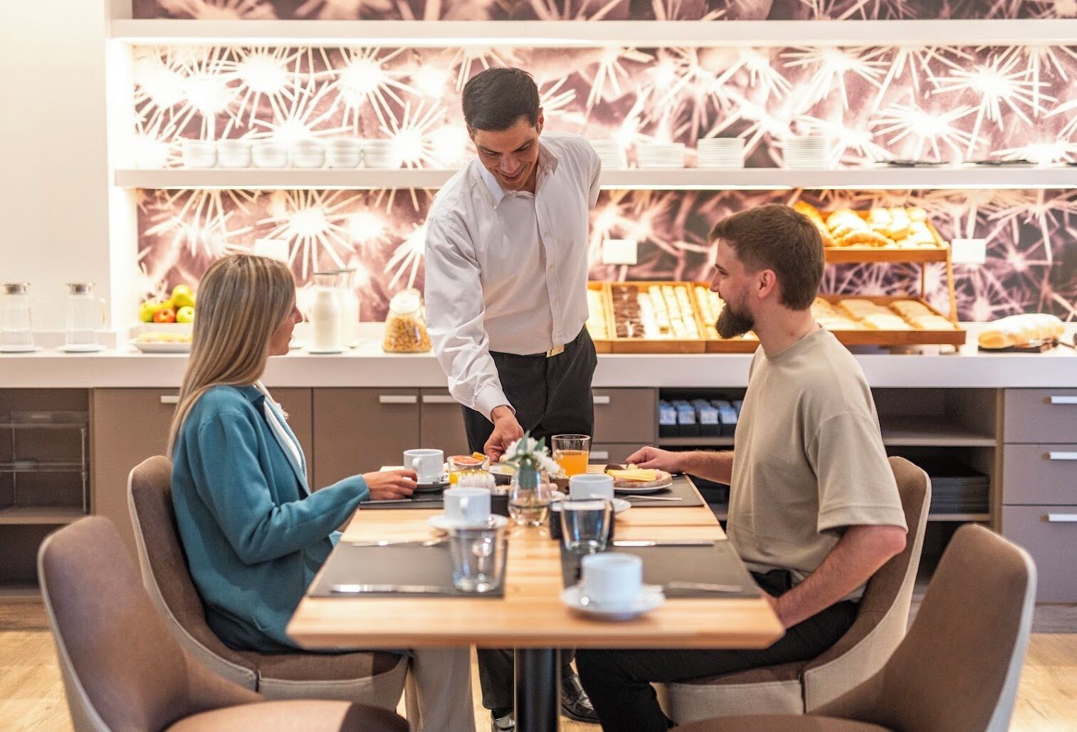 Disposição para o pequeno-almoço, com um empregado a servir comida, um buffet de pastelaria e uma parede decorativa com brilhos.