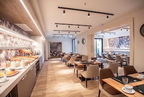 A modern dining area with a buffet on the left, multiple tables and chairs arranged for dining, and a large window providing natural light. - Dazzler By Wyndham Salta (Salta)