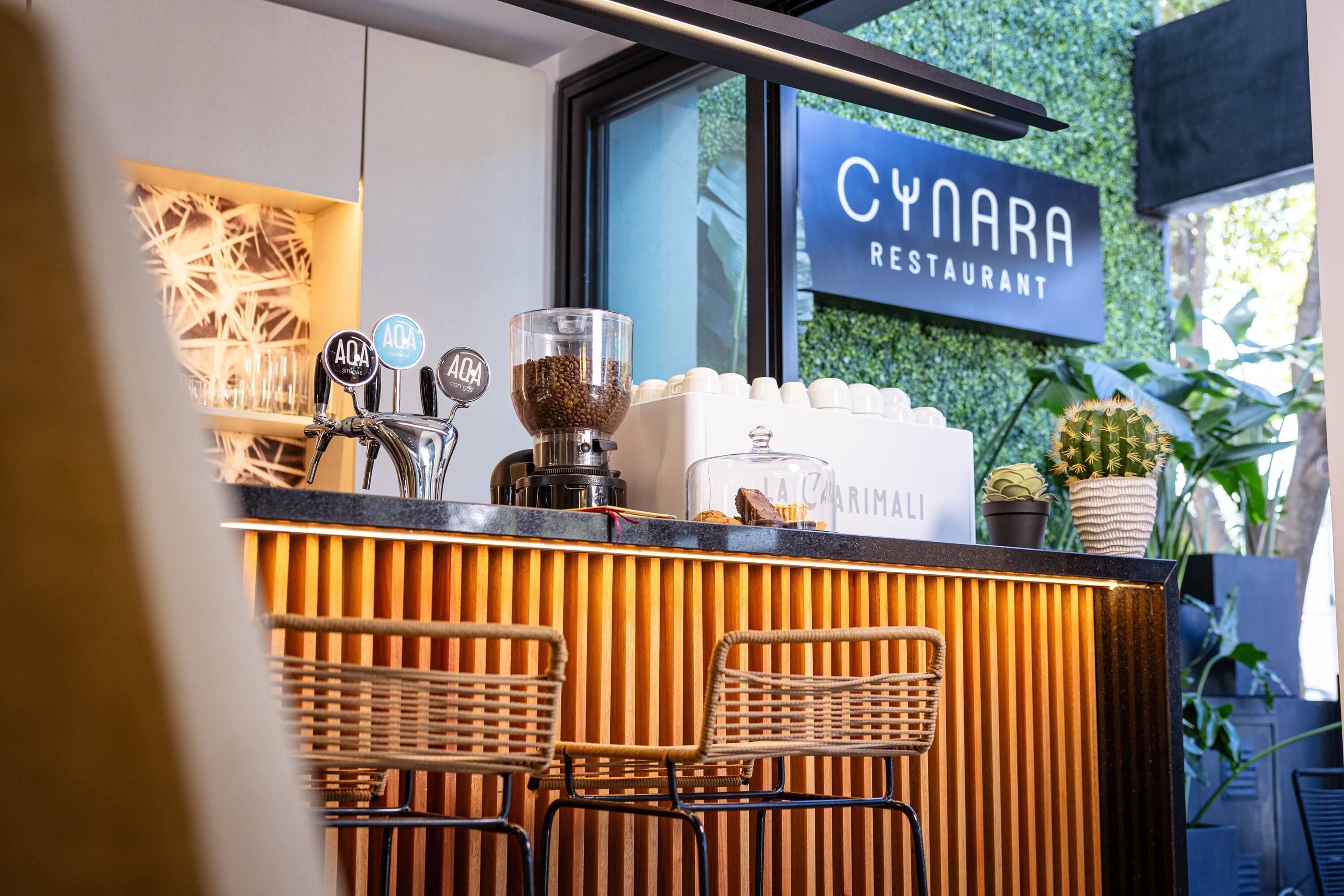 A café counter with a coffee machine, a glass container of coffee beans, and a sign for 'CYNARA RESTAURANT'.