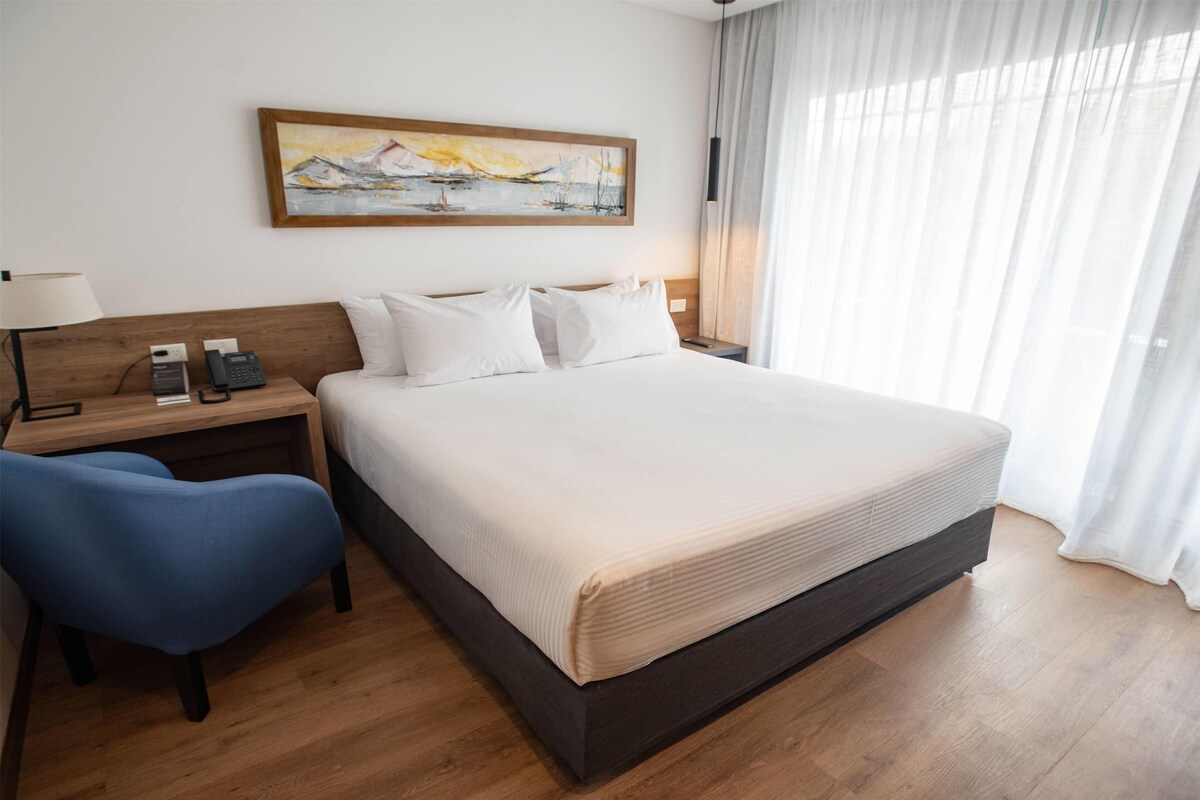 Una habitación de hotel con una cama grande, un escritorio con lámpara, una silla y un cuadro en la pared.