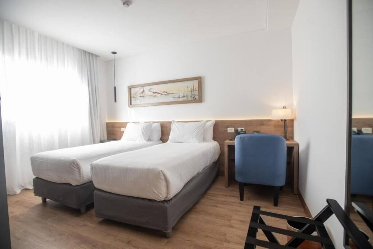 Een hotelkamer met twee bedden, een bureau, een stoel, een schilderij aan de muur en een raam met gordijnen.