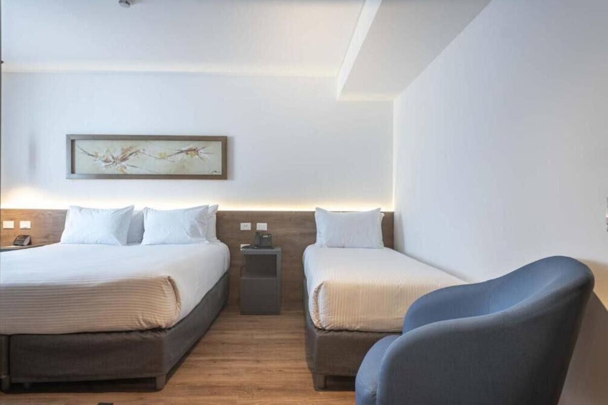 Een hotelkamer met twee bedden, een klein nachtkastje met een lamp, een lijst met een afbeelding aan de muur en een blauwe stoel.