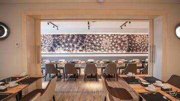 Un moderno salón de conferencias con varias mesas y sillas, un papel tapiz decorativo y vistas hacia un comedor.