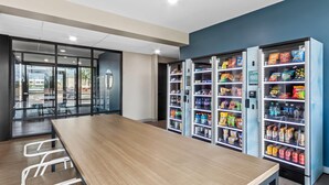 Un espace intérieur moderne, doté d’une longue table en bois et de chaises blanches, entouré de distributeurs automatiques remplis de collations et de boissons.