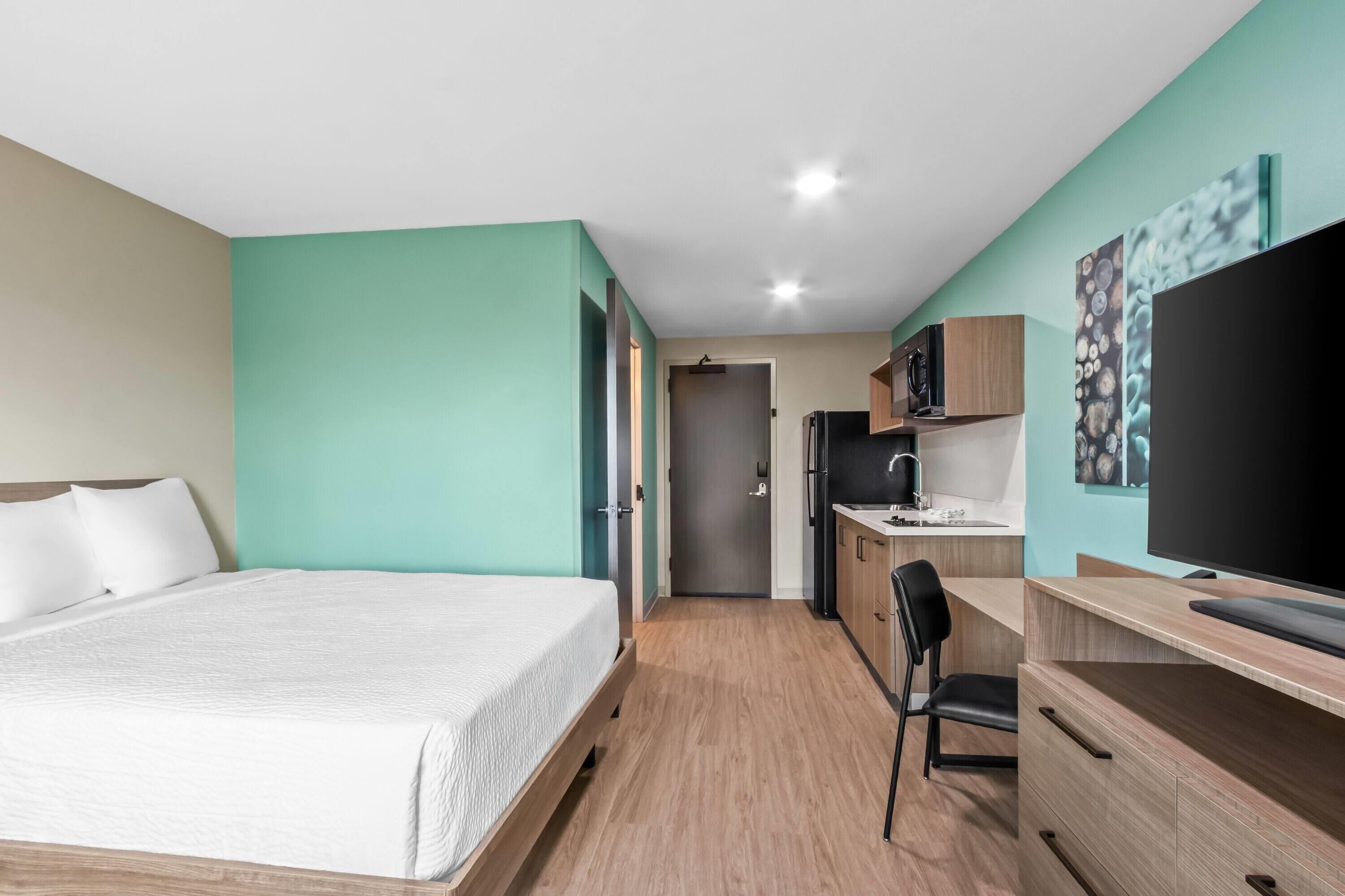 Habitación de hotel con cama, escritorio, silla, televisor y kitchenette.