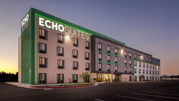 Il s’agit d’un hôtel moderne dont le nom, « Echo Suites », est affiché en grand sur la façade. Le bâtiment est composé de plusieurs étages, doté d’un toit plat et de rangées de fenêtres à chaque niveau.