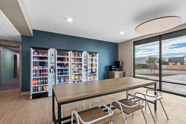 Un espacio interior moderno con una larga mesa de madera y sillas metálicas, rodeado de máquinas expendedoras llenas de aperitivos y bebidas.