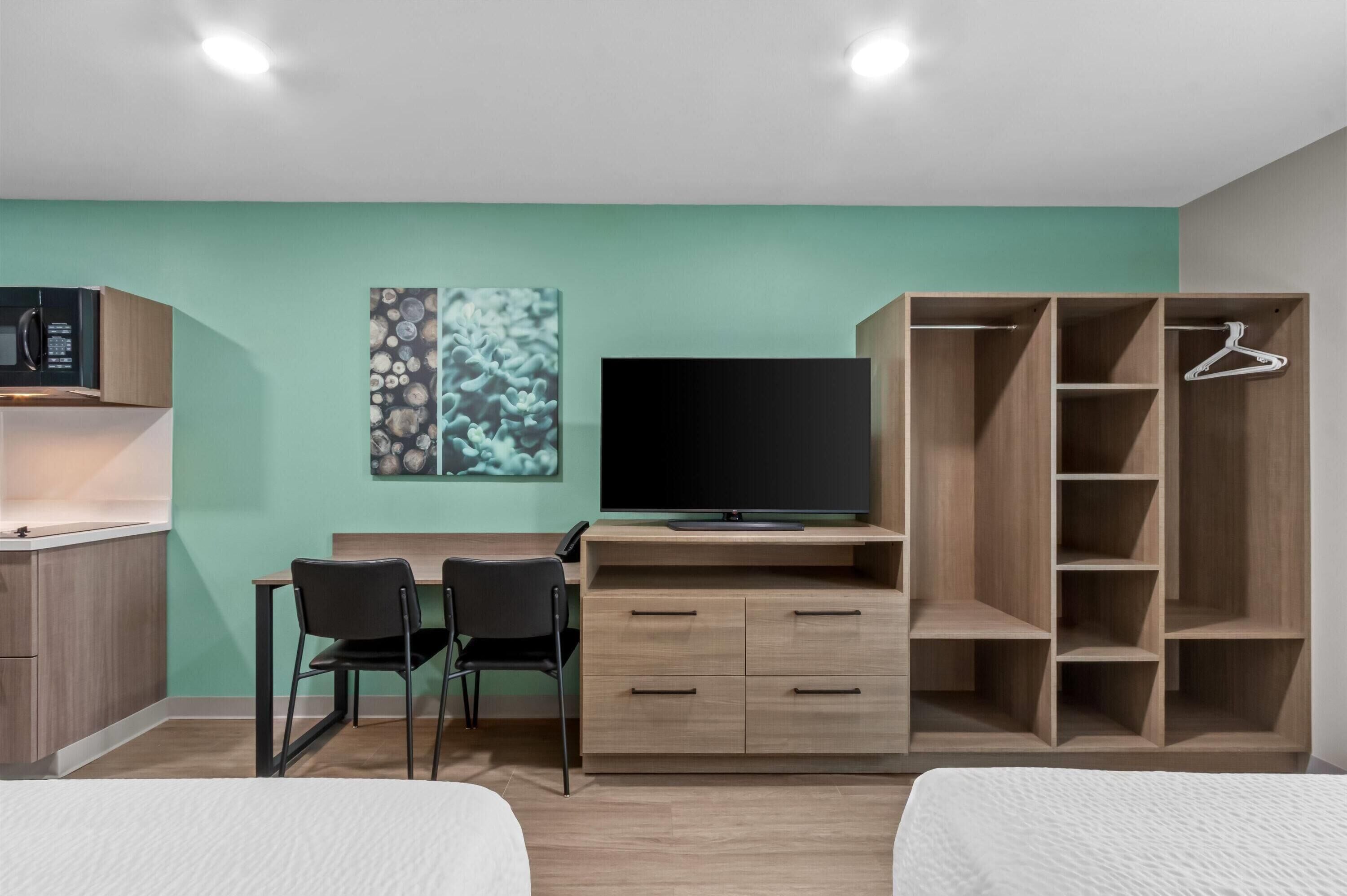 A modern hotel room with a kitchenette, a desk with two chairs, a television, and a wardrobe with multiple shelves.