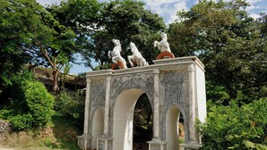 Une arche en pierre surmontée de trois statues de chevaux, le tout entouré de verdure et d’arbres.