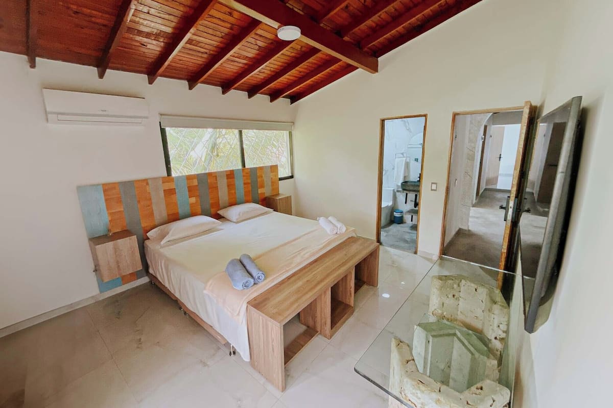 A bedroom with a wooden ceiling, a bed with white linens, a bench, a TV, and a window.
