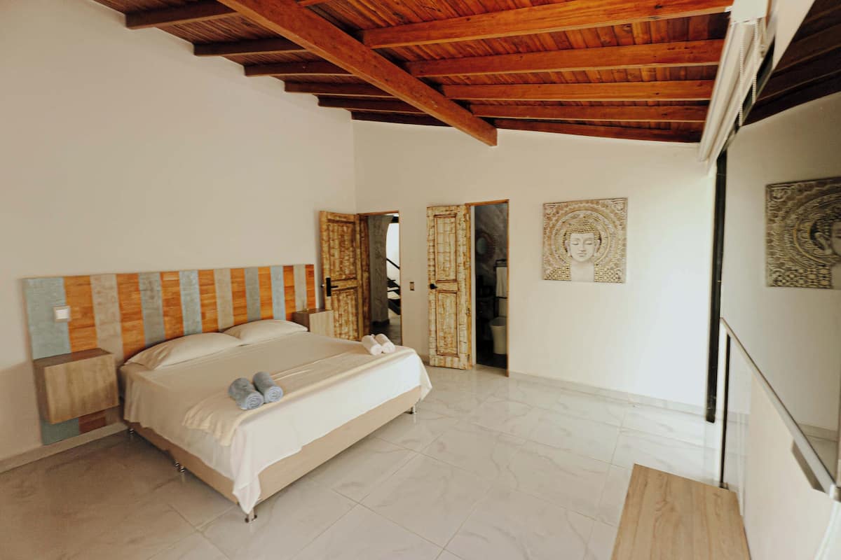 A bedroom with a wooden ceiling, a bed with a striped headboard, a large mirror, and a Buddha statue on the wall.