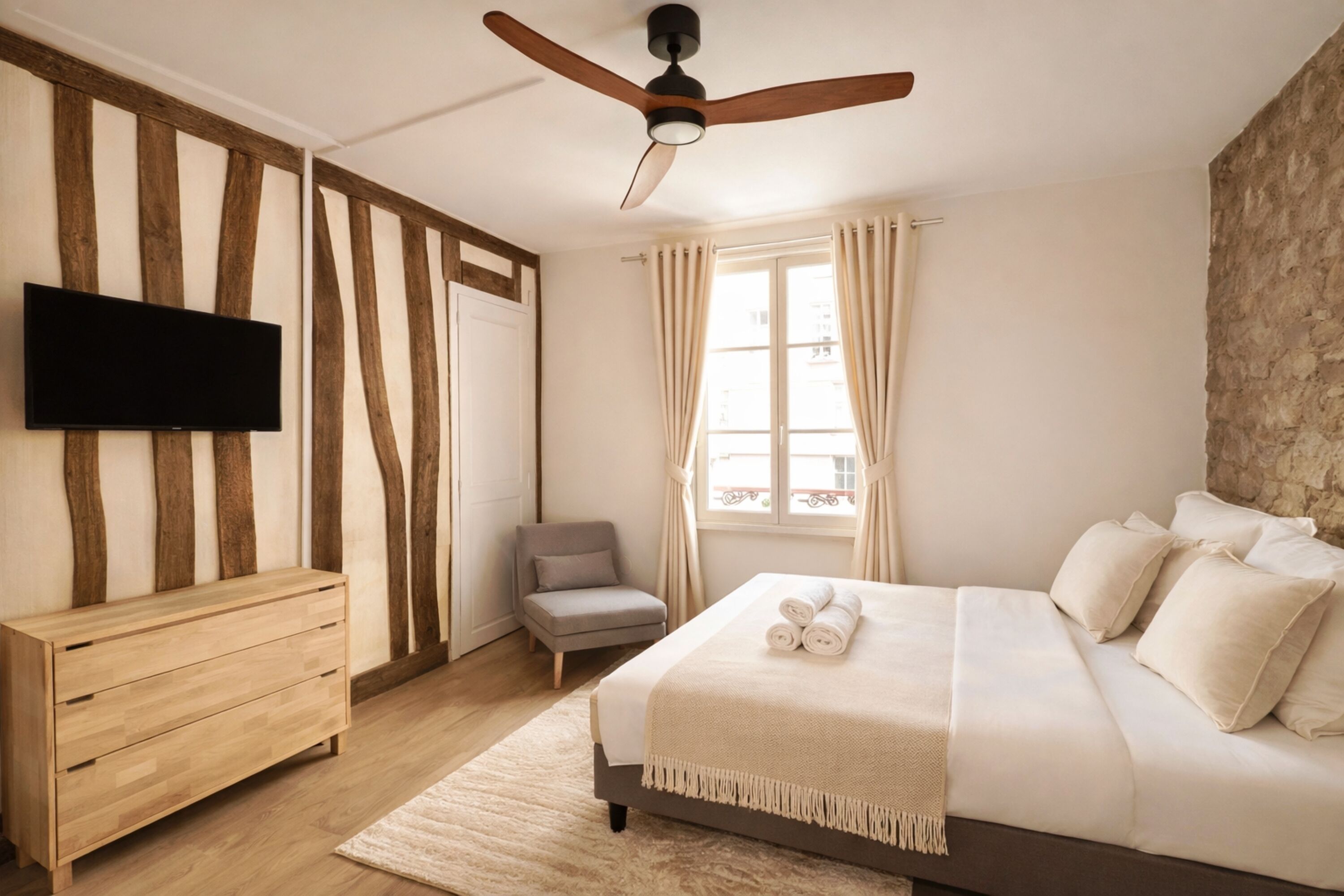 A bedroom with a bed, a TV, a ceiling fan, a chair, and a window with curtains.