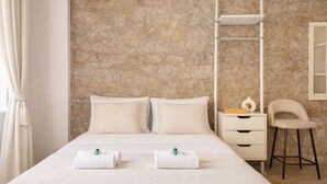 A neatly made bed with two towels, a nightstand with a book and a bottle, a chair, and a stone wall. - 1614 Sevres (Paris)