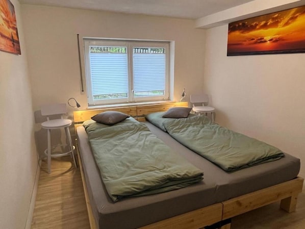 A small, single-room accommodation with two beds, a window with blinds, a desk, and a chair.