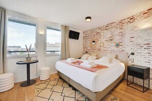 Une chambre d’hôtel moderne avec un grand lit, un mur décoratif au motif de briques, une petite table ronde et une vue sur la ville.