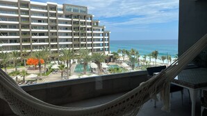 A balcony with a hammock, overlooking a resort area with a pool, palm trees, and a view of the ocean. - Cabo San Lucas! - It's your choice - Jr. Suite, 1 or 2-bedroom resort stay! (Cabo San Lucas)