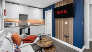 A modern kitchen with white cabinets, a microwave, and a coffee maker. A large TV mounted on the wall displays "NETFLIX".