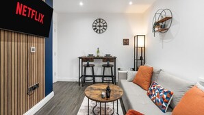 Una sala de estar moderna con un cartel de Netflix, una mesa de centro redonda, un sofá con cojines naranjas, un reloj de pared y una pequeña zona de comedor con una mesa y sillas.