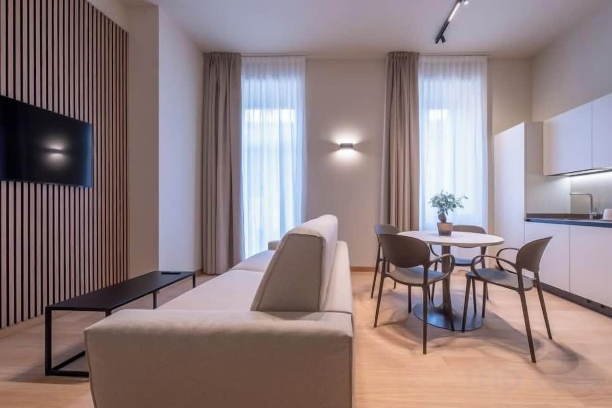 A modern hotel room with a sofa, dining table, chairs, a flat-screen TV, and a kitchen area.