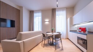 A modern living room with a sofa, a dining table with chairs, a kitchen area with a red appliance, and a flat-screen TV mounted on a wooden panel. - Oriana Homèl Milano (Milan)