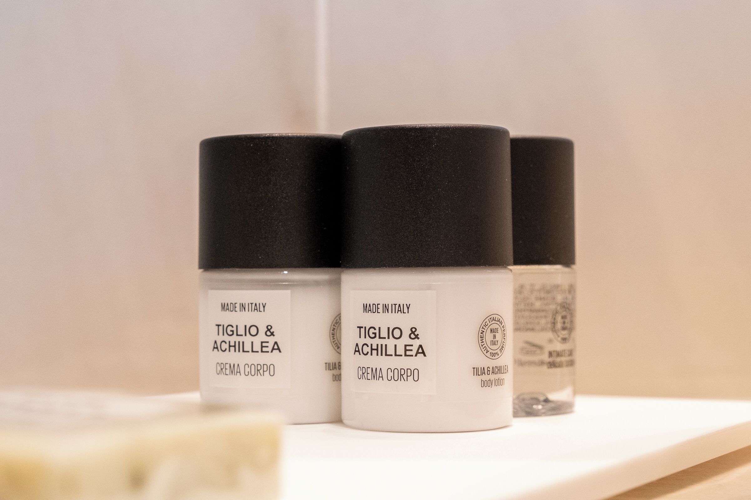 Three white containers with black lids labeled "Tiglio & Achillea Crema Corpo" on a white shelf.