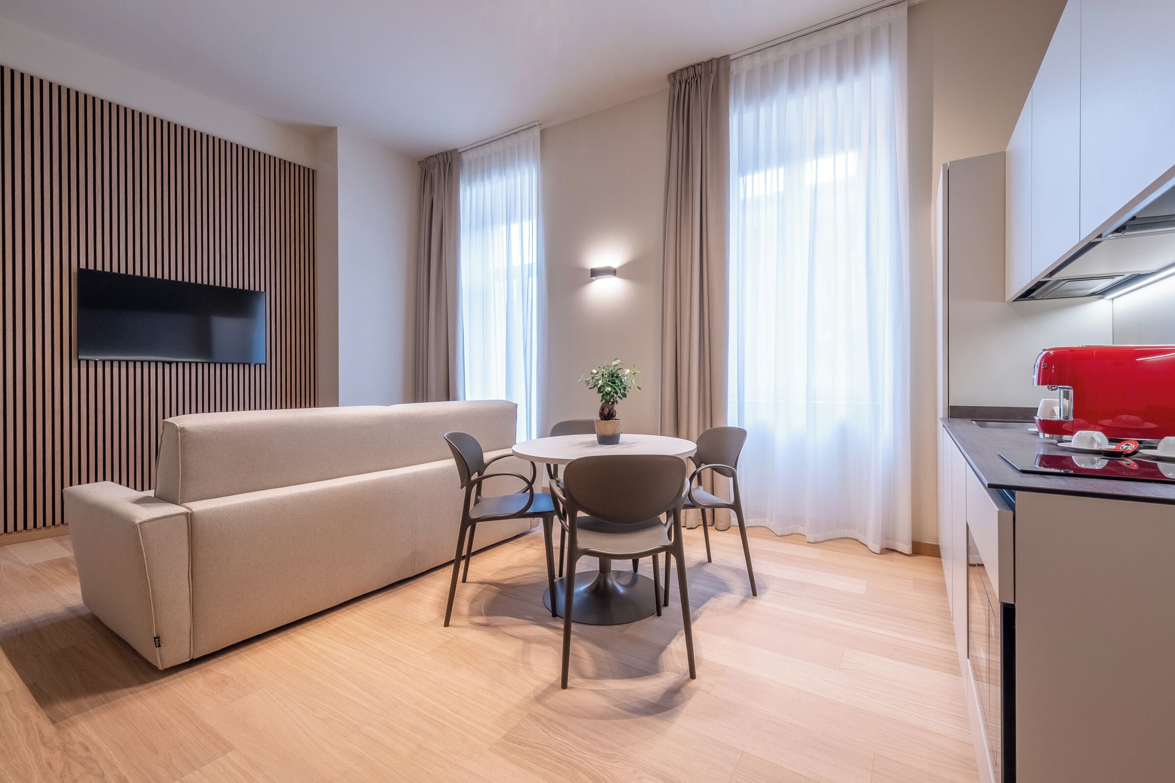 A modern living room with a sofa, a dining table with chairs, a flat-screen TV mounted on a wooden panel, and a kitchen area with a red appliance.