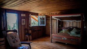 A wooden cabin interior with a four-poster bed, a chair with a cushion, a small table, and a window with a view of trees.