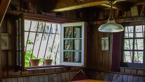 A rustic wooden interior with a large table and benches, large windows with potted plants, and a hanging light fixture.
