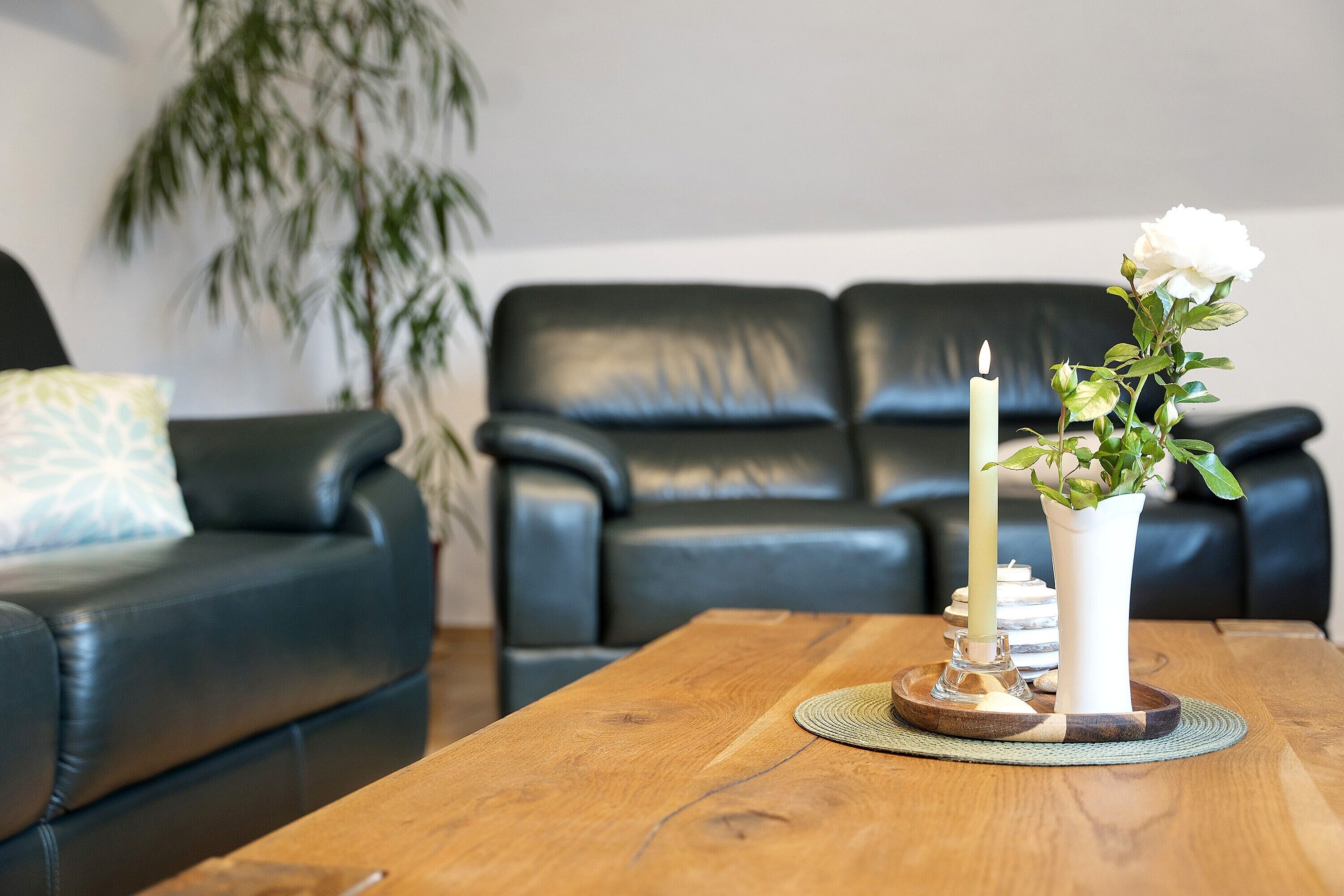 Een woonkamer met een houten salontafel, een zwarte leren bank, een witte vaas met een bloem en een brandende kaars.