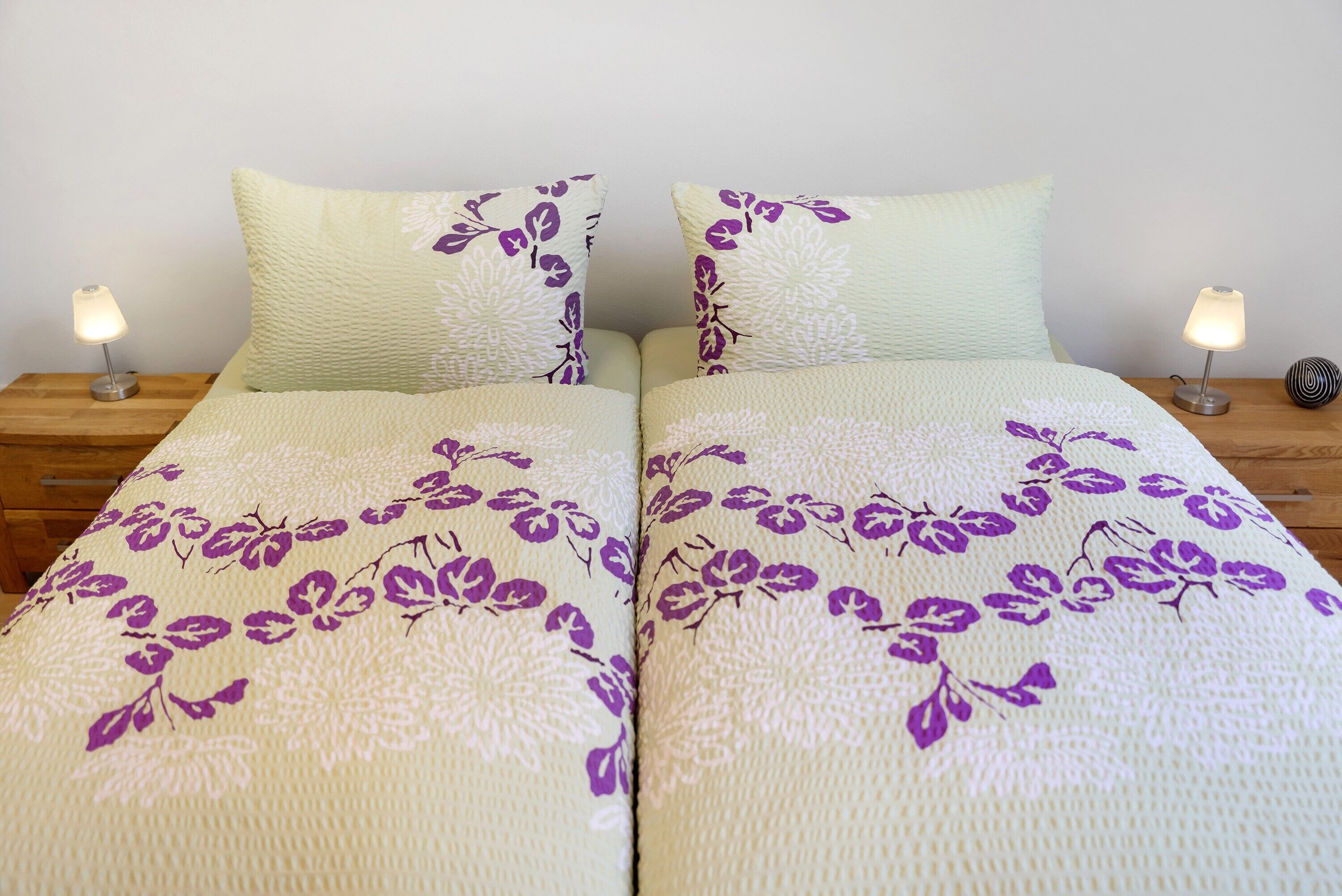 Uma cama de casal com roupa de cama estampada em flor, duas luminárias de cabeceira e uma mesinha de cabeceira de madeira.
