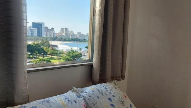 A bedroom with a bed, a window with curtains, and a view of a cityscape and beach. - SUH Botafogo 03 Great Bars and Shopping (Rio de Janeiro)