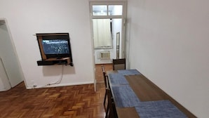 A room with a wooden herringbone floor, a television mounted on the wall, a dining table with blue placemats, and a door leading to another room. - SUH Botafogo 03 Great Bars and Shopping (Rio de Janeiro)