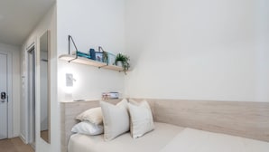Ein Schlafzimmer mit Bett, Kissen, einem Nachttisch mit Lampe, einem Regal mit Büchern und einer Pflanze sowie einer Tür.