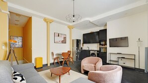 Een moderne woonkamer met een grijze bank, een roze fauteuil en een houten salontafel. In de achtergrond is een keuken met donkere kastjes en een televisie te zien.