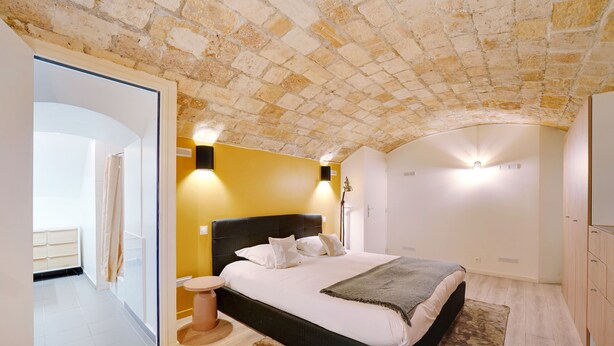 Een slaapkamer met een bakstenen plafond, een bed met witte beddengoed, een rond bijzettafeltje en een houten garderobe.