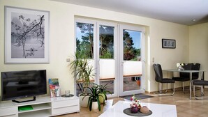 Un soggiorno con televisione, un tavolo da pranzo con sedie, una porta scorrevole in vetro che dà su un balcone e un quadro appeso al muro.