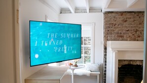 Habitació amb televisió que mostra "THE SUMMER I TURNED PRETTY" sobre un fons color turquesa, una llar de foc blanca i una paret de pedra.
