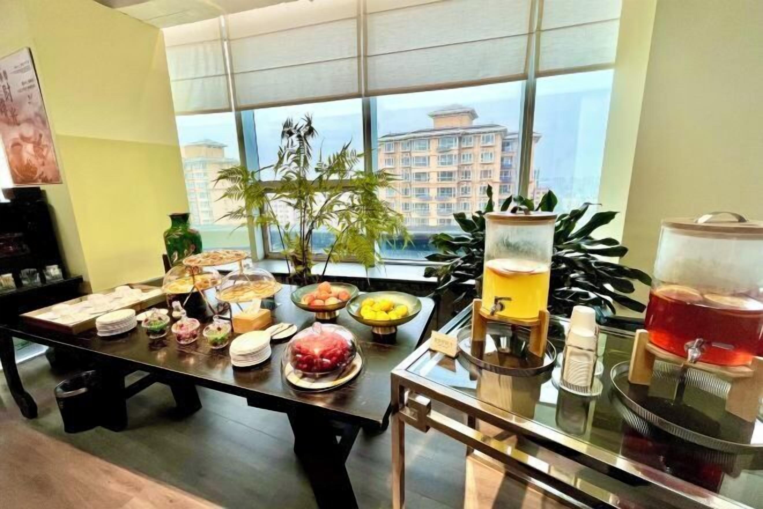 A breakfast buffet with various food items and beverages, a large window with a view of a building, and a potted plant on the table.