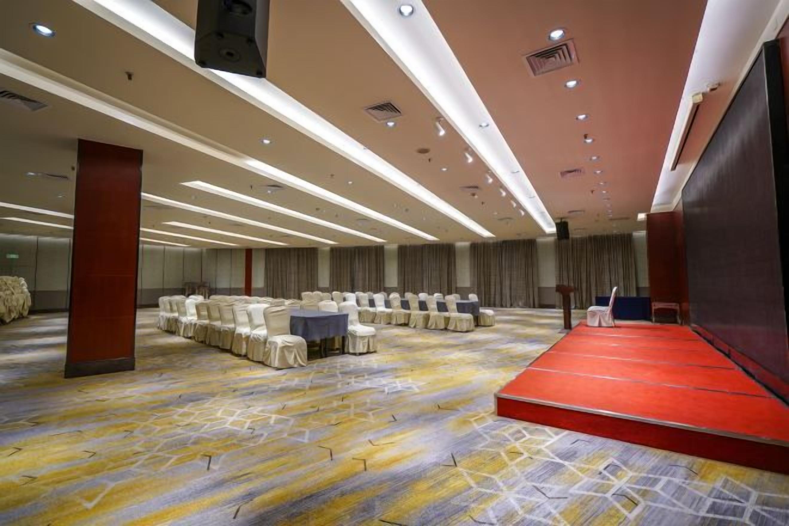 A conference room with rows of tables and chairs, a stage, and a carpeted floor.