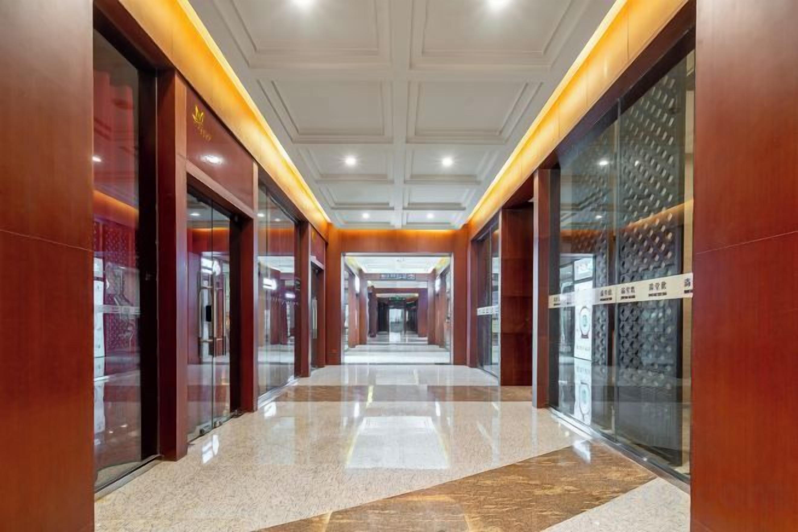 A corridor with glass doors on the left, a polished floor, and a ceiling with recessed lighting.