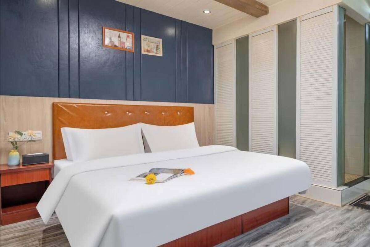 A hotel room with a wooden headboard, a white bedspread, a nightstand with a vase, and a large window with blinds.