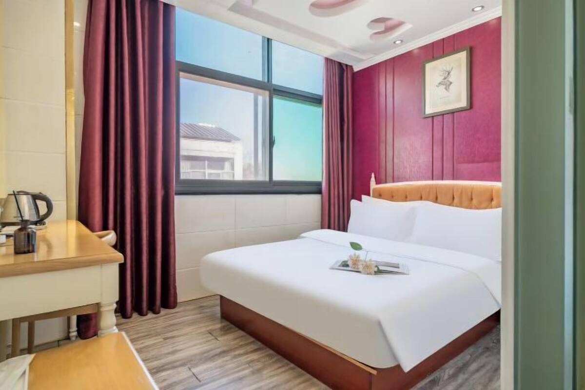 A hotel room with a bed, a desk, a window with curtains, and a wall-mounted picture.