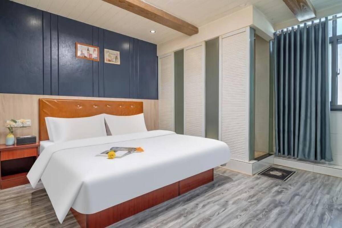 A modern hotel room with a wooden headboard bed, a nightstand with a lamp, a large window with blinds, and a wooden floor.