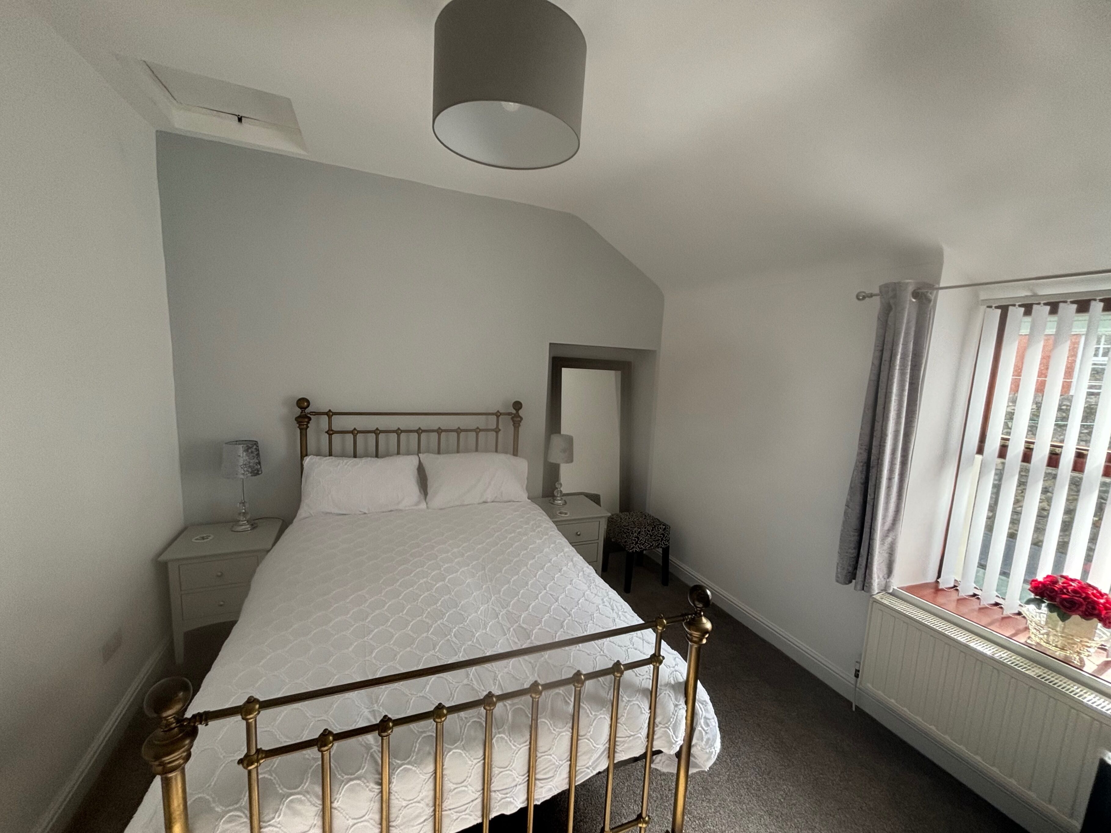 Une chambre à coucher avec un lit, des tables de chevet, une lampe, une fenêtre avec des rideaux et un radiateur.