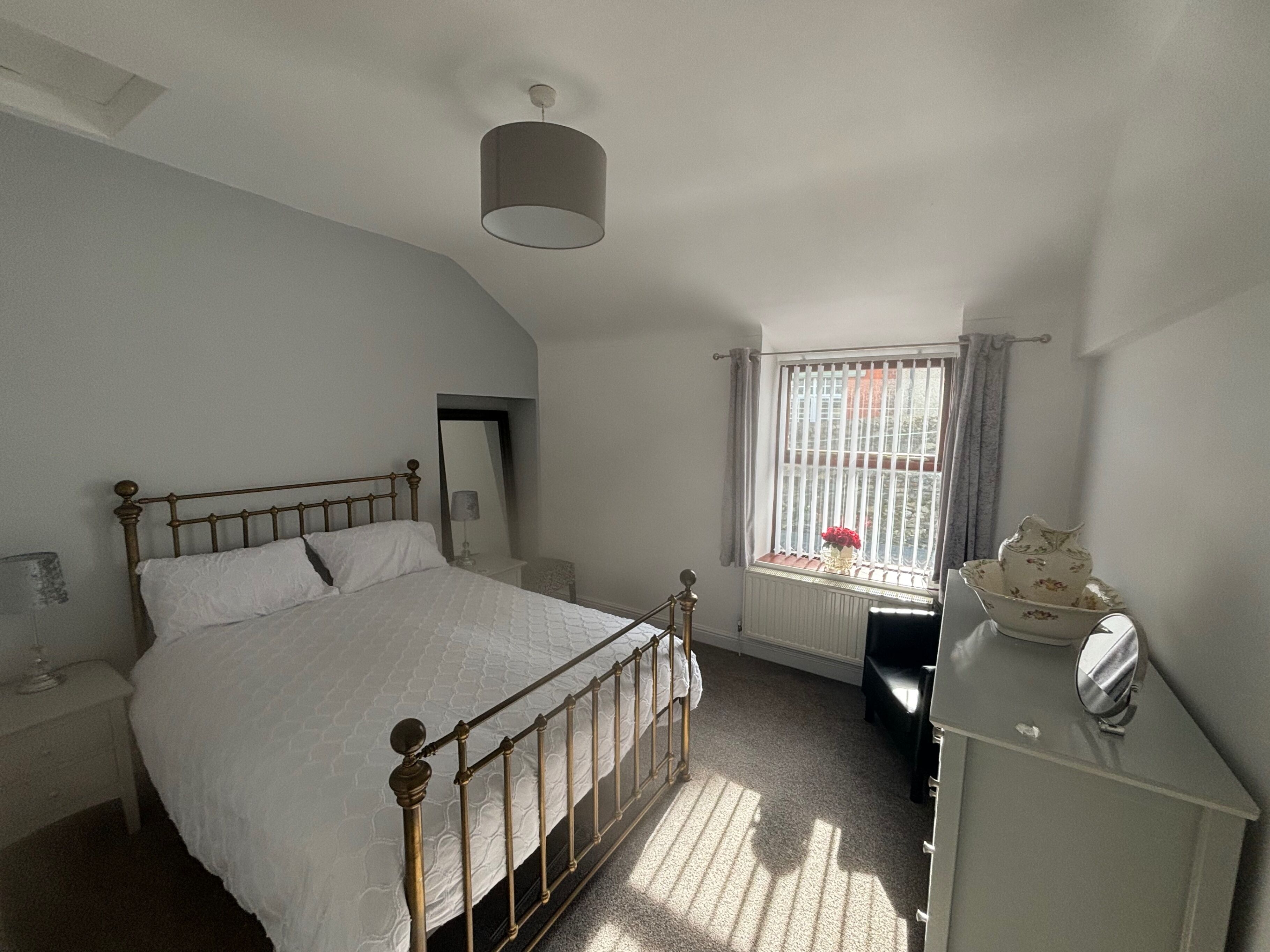 A bedroom with a bed, a nightstand, a dresser, a mirror, a window with curtains, and a radiator.